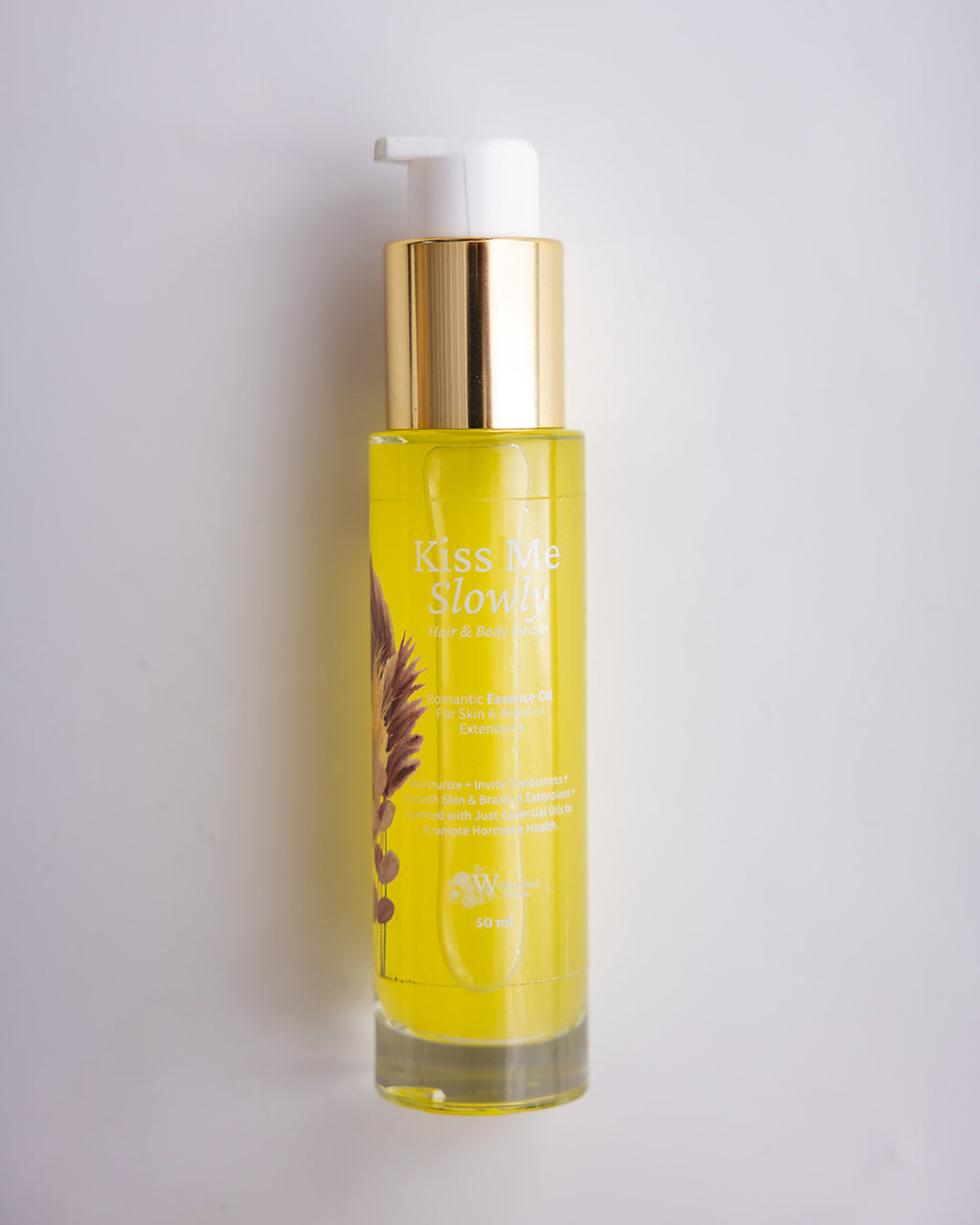 A bottle of Kiss Me Slowly hair and body  oil with a white pump and gold collar, filled with yellow oil, set against a plain light gray background.