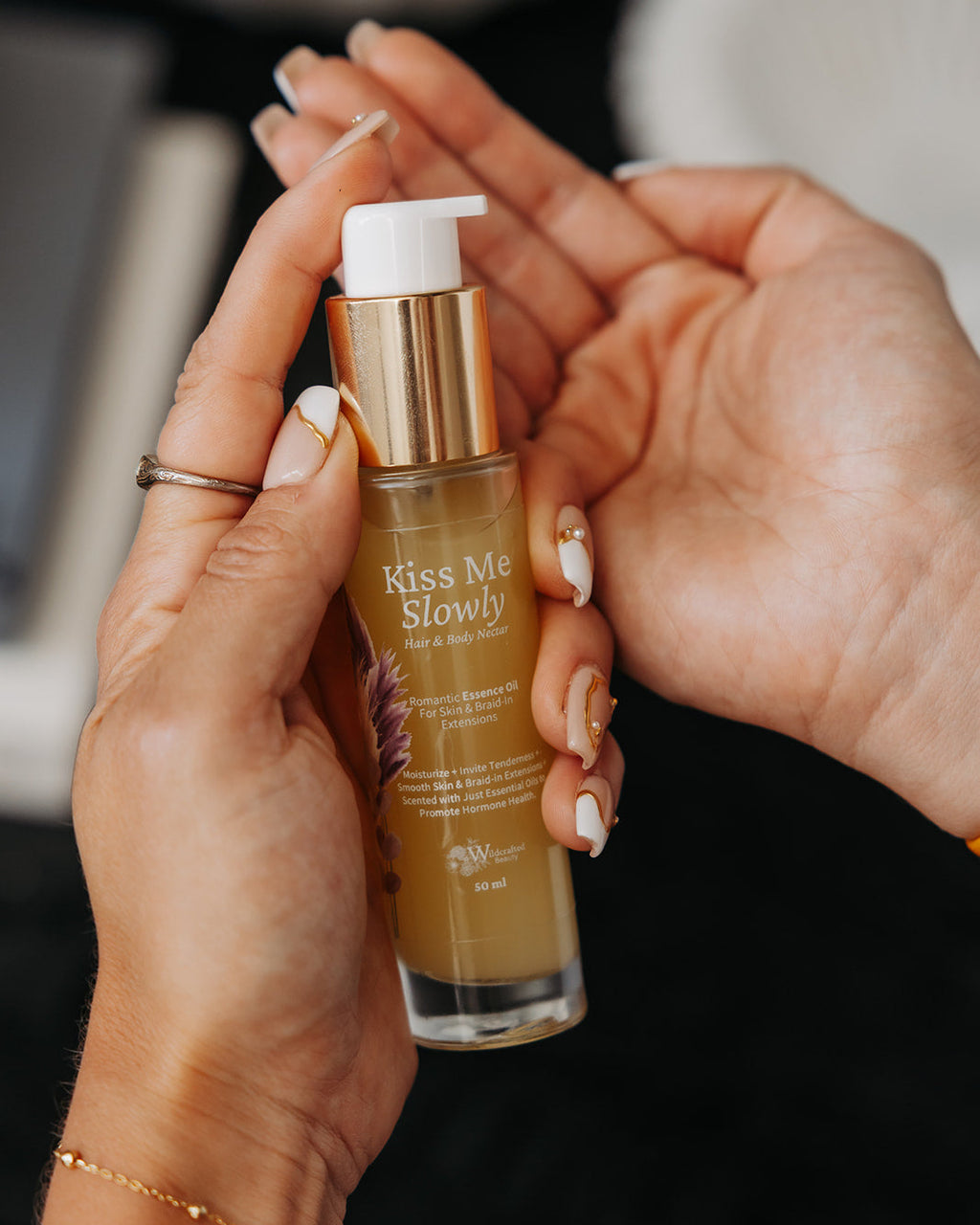 A person holds a bottle of 'Kiss Me Slowly' hand cream. The body oil bottle is clear glass showing a golden-yellow oil inside, with a metallic gold pump and a white pump cap.