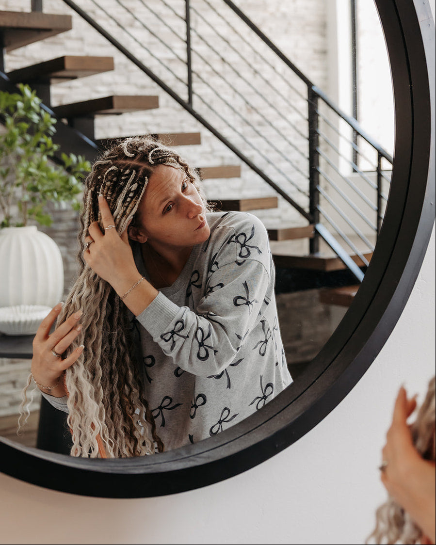 A woman with long, light brown and blonde box braids looks into a large round wall mirror. She is wearing a light gray long-sleeve shirt with a black graphic pattern.