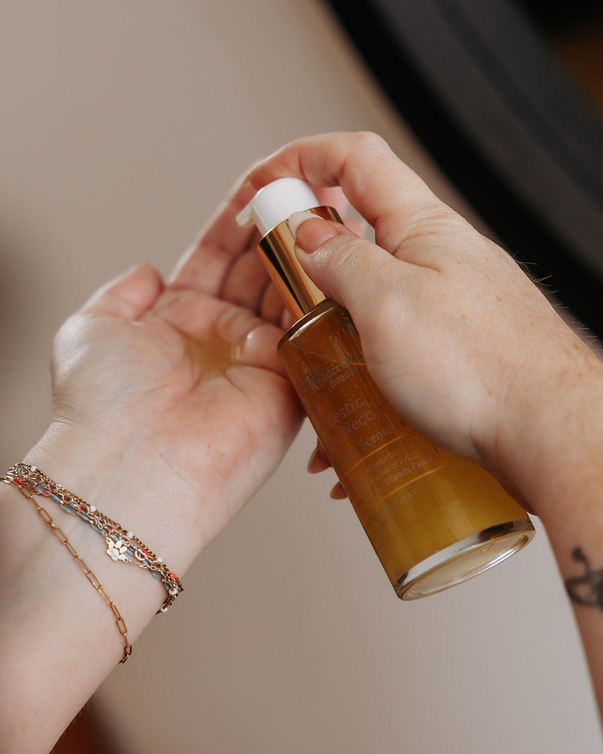 A close-up of a person's hands holding a clear bottle filled with golden amber liquid, with one hand receiving the liquid and the other holding the pump. The person is wearing delicate rose gold bracelets.