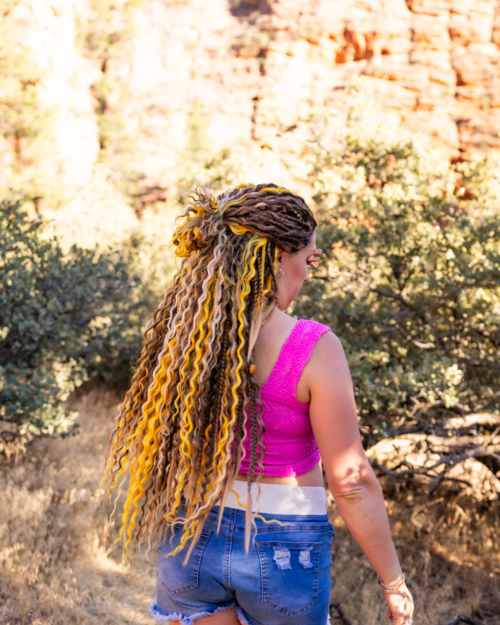 A woman standing outdoors in a desert canyon setting, viewed from behind, wearing a bright pink crop top and denim shorts. She has long, curly dreadlocks with prominent yellow and brown highlights.