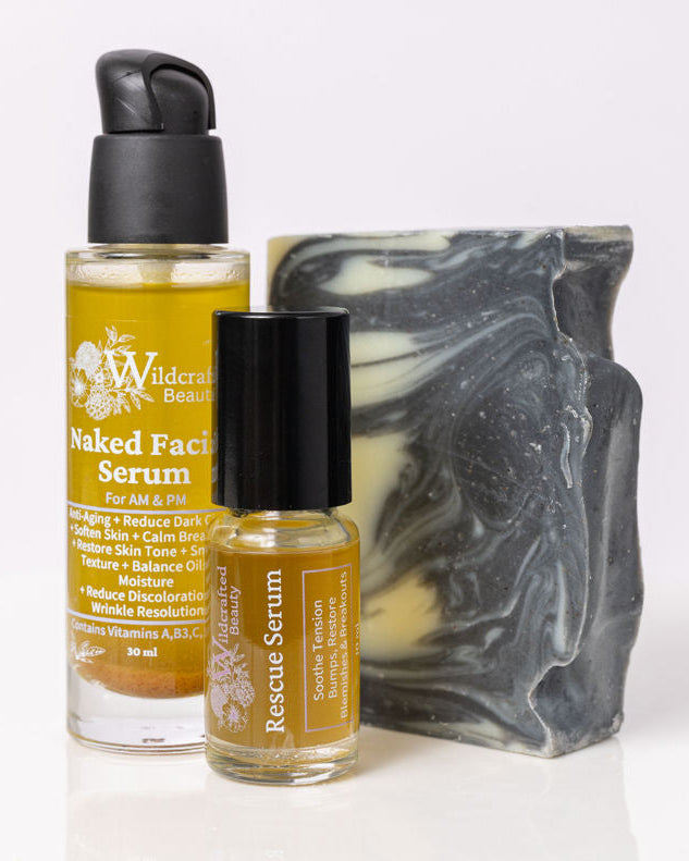 A set of skincare products including a black and white swirled Facial Cleansing Bar, a yellow pump bottle labeled Naked Facial Serum, and a small roller bottle labeled Rescue Serum, all placed on a white surface.