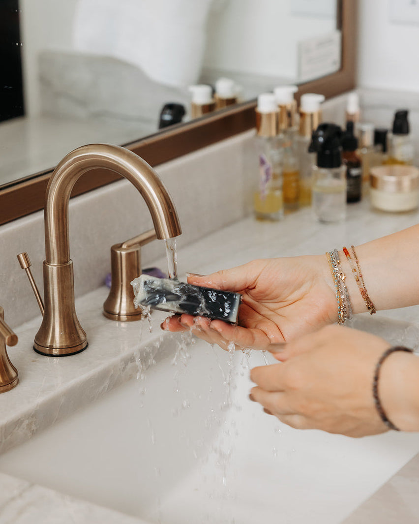 A person's hands holding a black and white marbled facial bar under running water from a matte bronze faucet in a bathroom sink, with toiletries visible in the background.