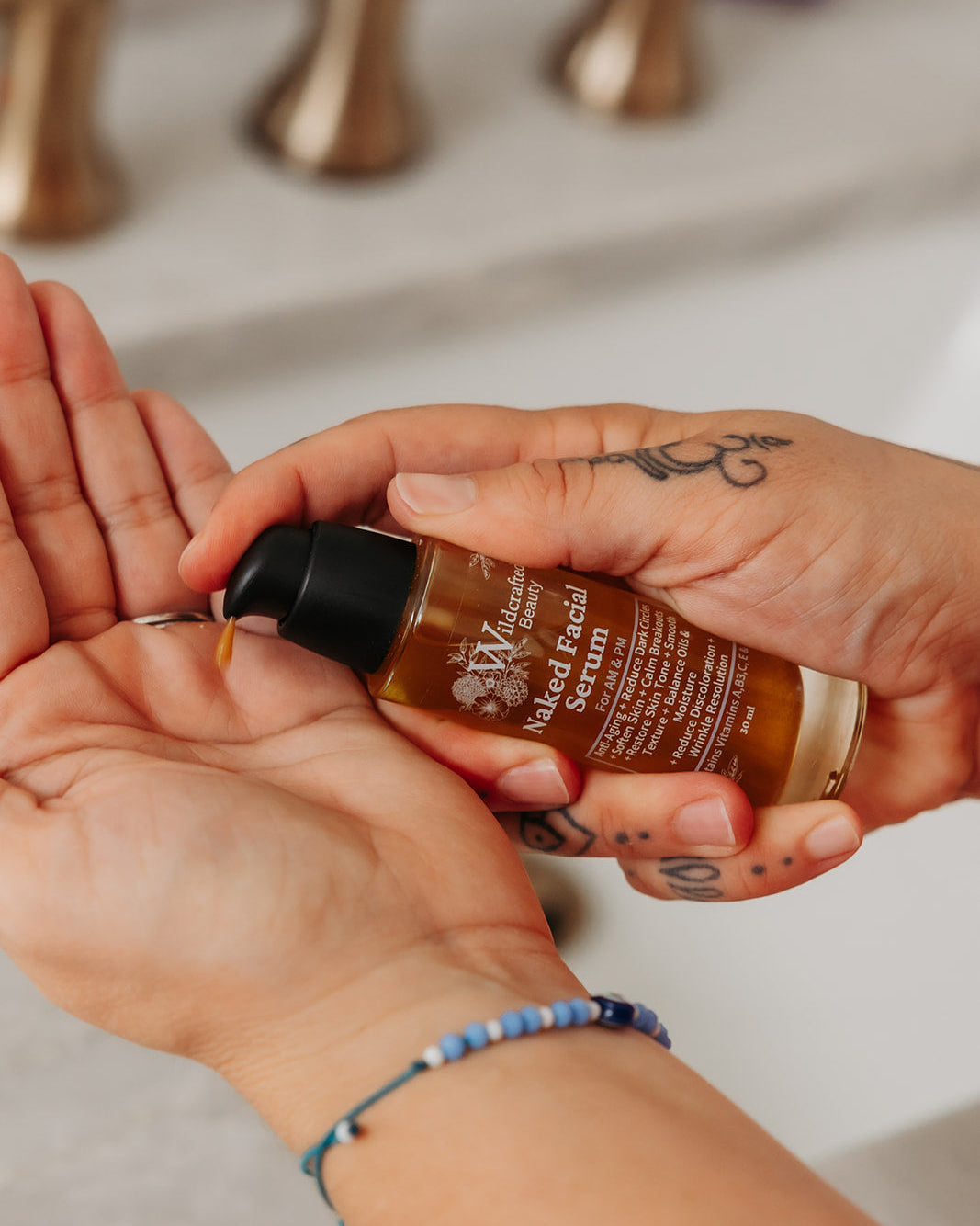 A close-up of a person's hands dispensing a golden-brown liquid from a small amber bottle labeled 'Naked Facial Serum' into their palm, with a blurred bathroom sink and faucet in the background.