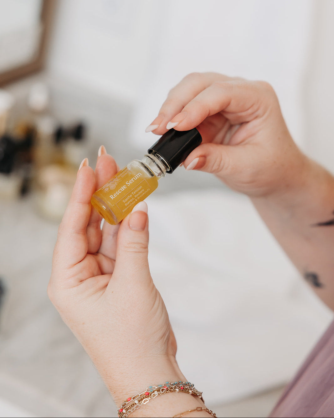 A close-up of a person's hands holding a small amber glass bottle with a black cap, showing the golden-yellow liquid inside. The background is a softly blurred bathroom or vanity setting with light-colored tiles and other bottles.