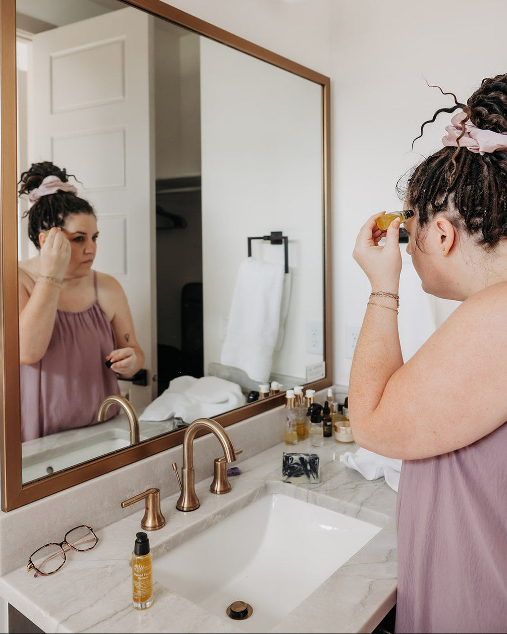 A woman with her hair in a messy bun applies Rescue Serum while looking into a large wood-framed bathroom mirror. She is wearing a mauve sleeveless top, and the bathroom features a white sink, brass faucet, and a light fixture above.