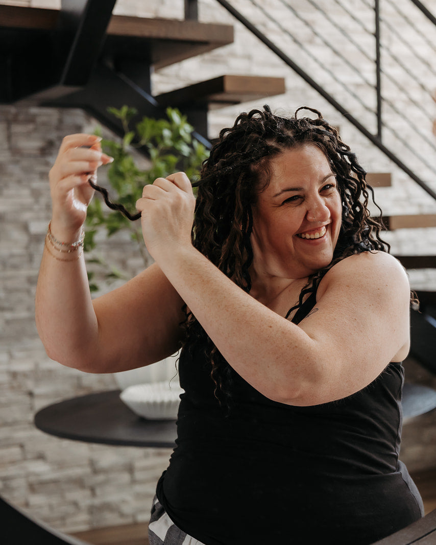A woman with short wavy dark hair extensions smiles while looking at her reflection in a large round mirror, separating the dreads. A staircase and indoor setting are visible in the background.