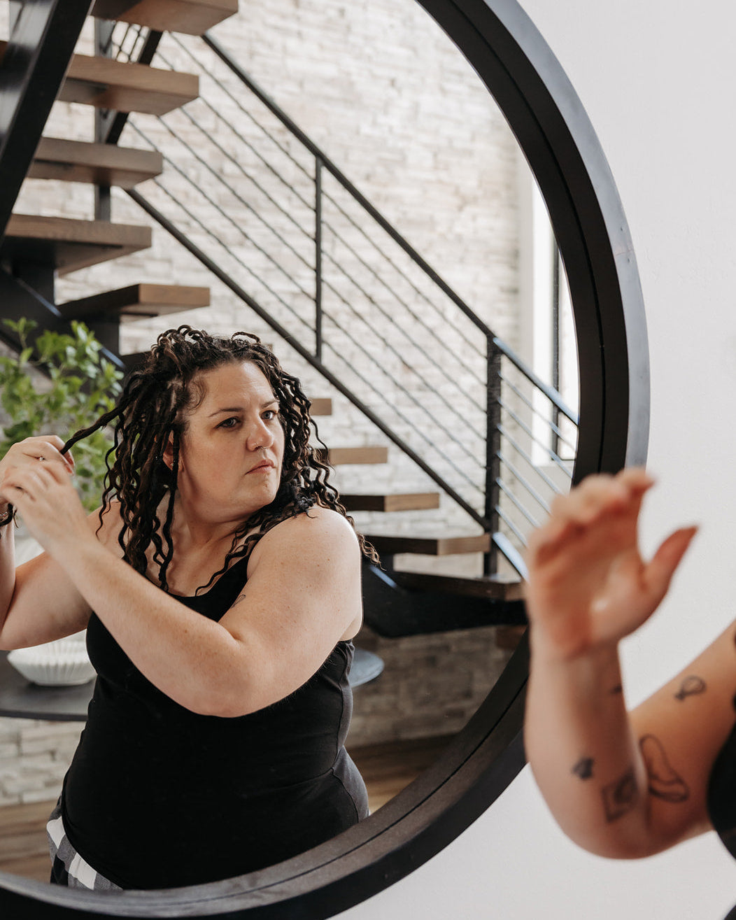 A woman with wavy dark braid-in hair extensions wearing a black tank top is seen in a large round mirror, holding a hairbrush near her hair. The reflection of her arm with a tattoo is visible in the foreground, and a modern staircase with wooden steps and black railings is in the background.