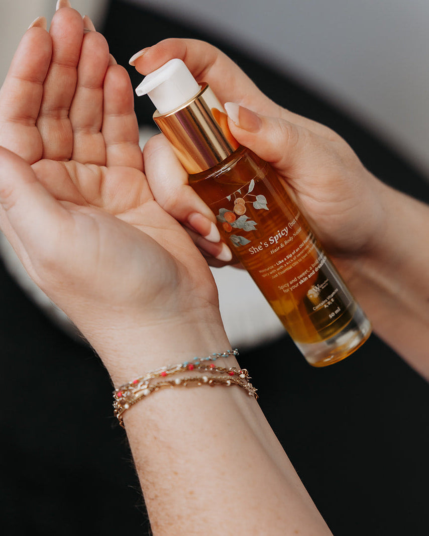 A person holding a bottle of amber-colored hair oil with a white pump dispenser, with some of the oil being dispensed onto their palm. The person has visible arm tattoos and a delicate bracelet on one wrist.