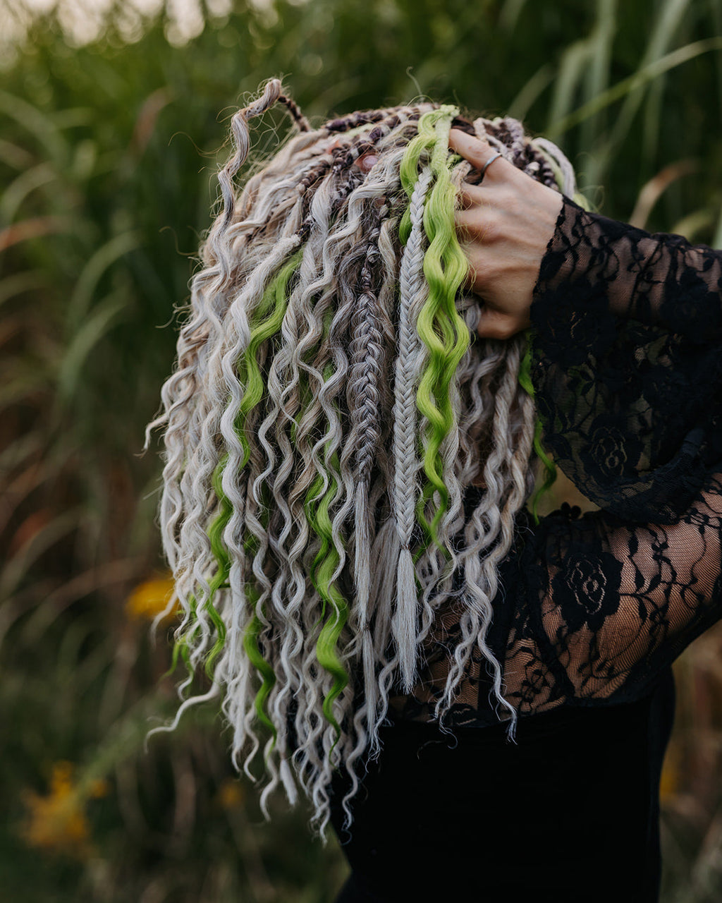 A person outdoors wearing a black lace top with her hands in her wavy and braided dreadlocks in white, gray, and bright lime green. Tall green grass is blurred in the background.