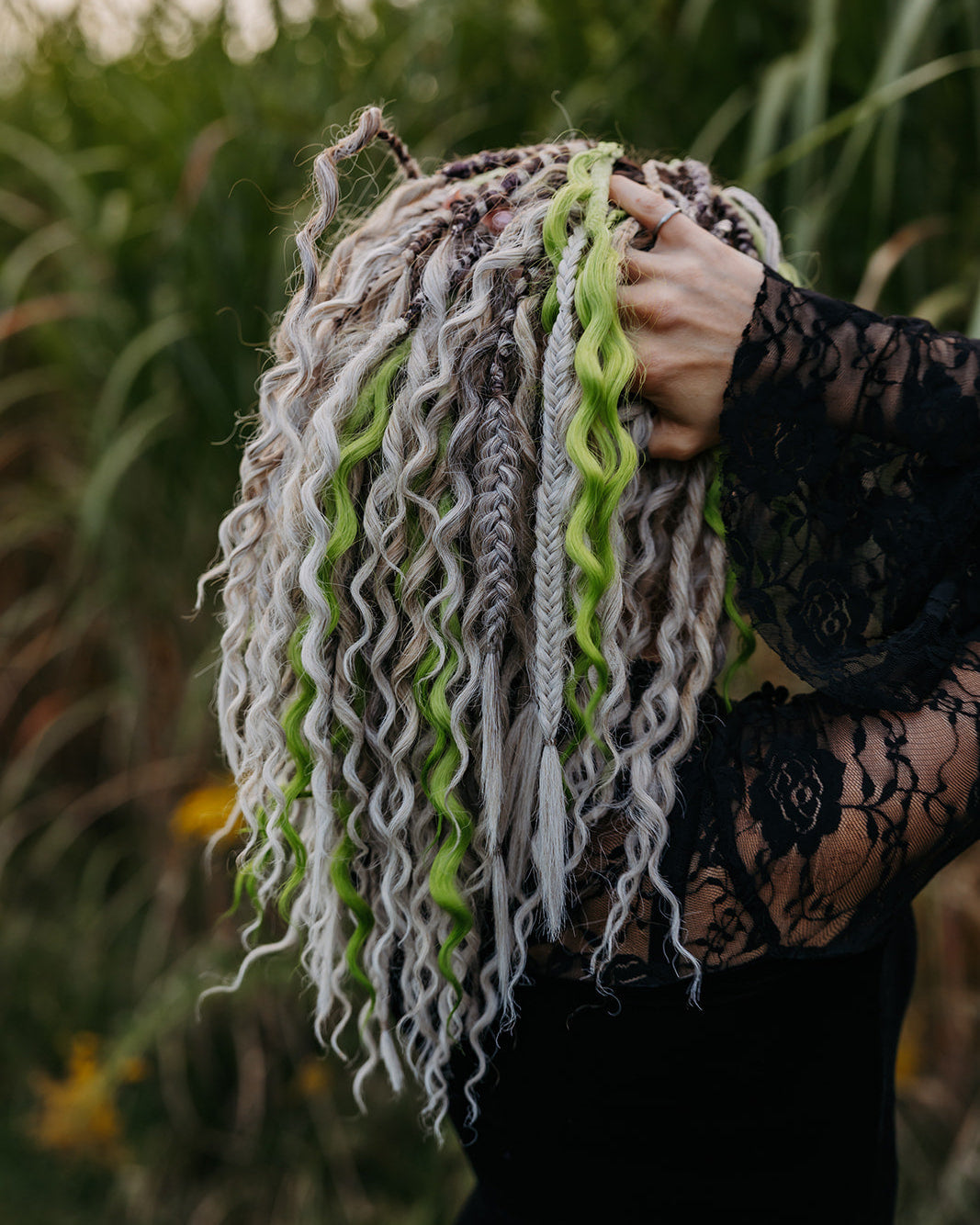 A person outdoors wearing a black lace top with her hands in her wavy and braided dreadlocks in white, gray, and bright lime green. Tall green grass is blurred in the background.