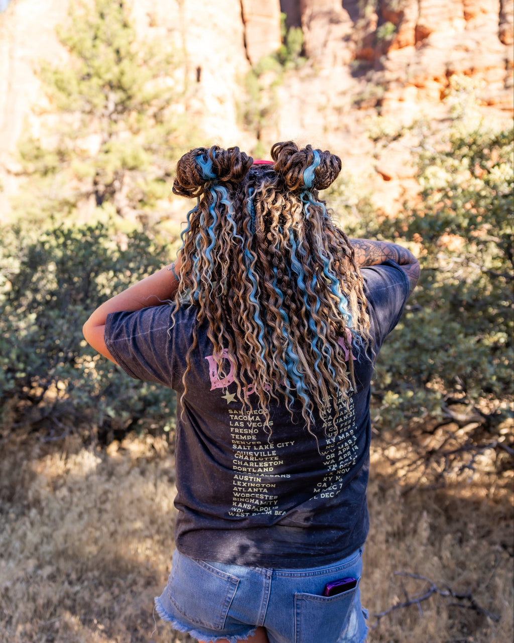 A person standing outdoors in a rocky, brushy landscape, photographed from behind. The person has curly dreadlocks styled in two top buns, wearing a dark graphic T-shirt and denim shorts.