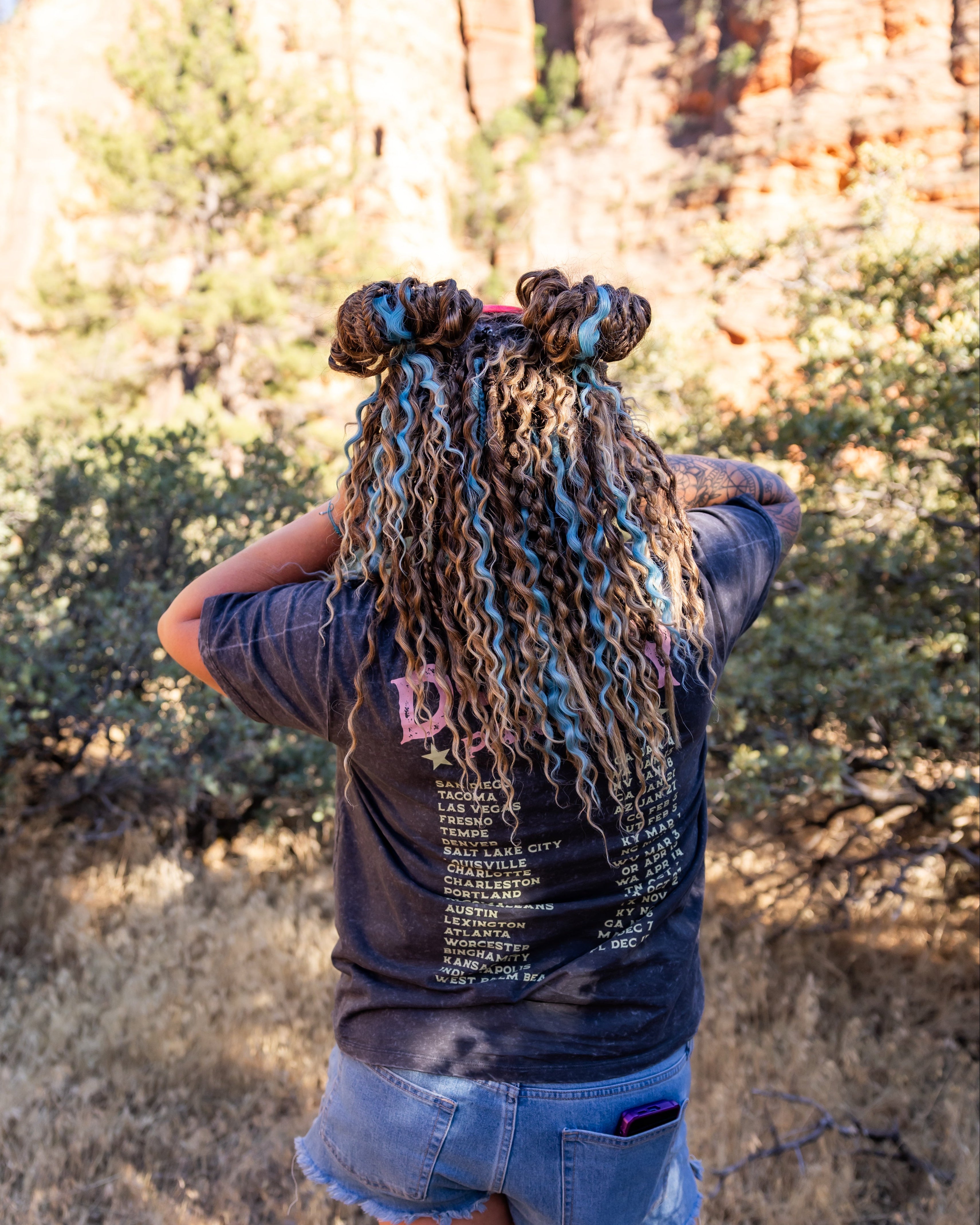 A person standing outdoors in a rocky, brushy landscape, photographed from behind. The person has curly dreadlocks styled in two top buns, wearing a dark graphic T-shirt and denim shorts.