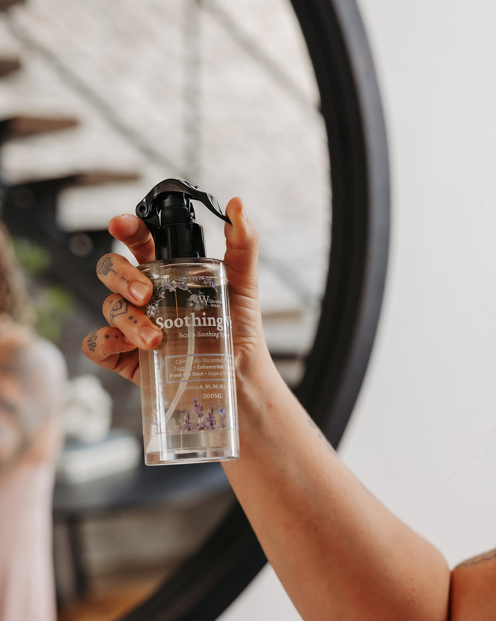 A hand holding a clear pump bottle labeled 'Soothing Veil' in front of a mirror with a blurred interior background.