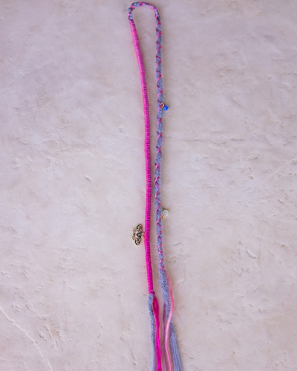 A handmade bohemian-style straight grey dread with pink braided wrap fiber, a small gold-toned butterfly charm, and colorful charms, laid flat on a textured off-white surface.