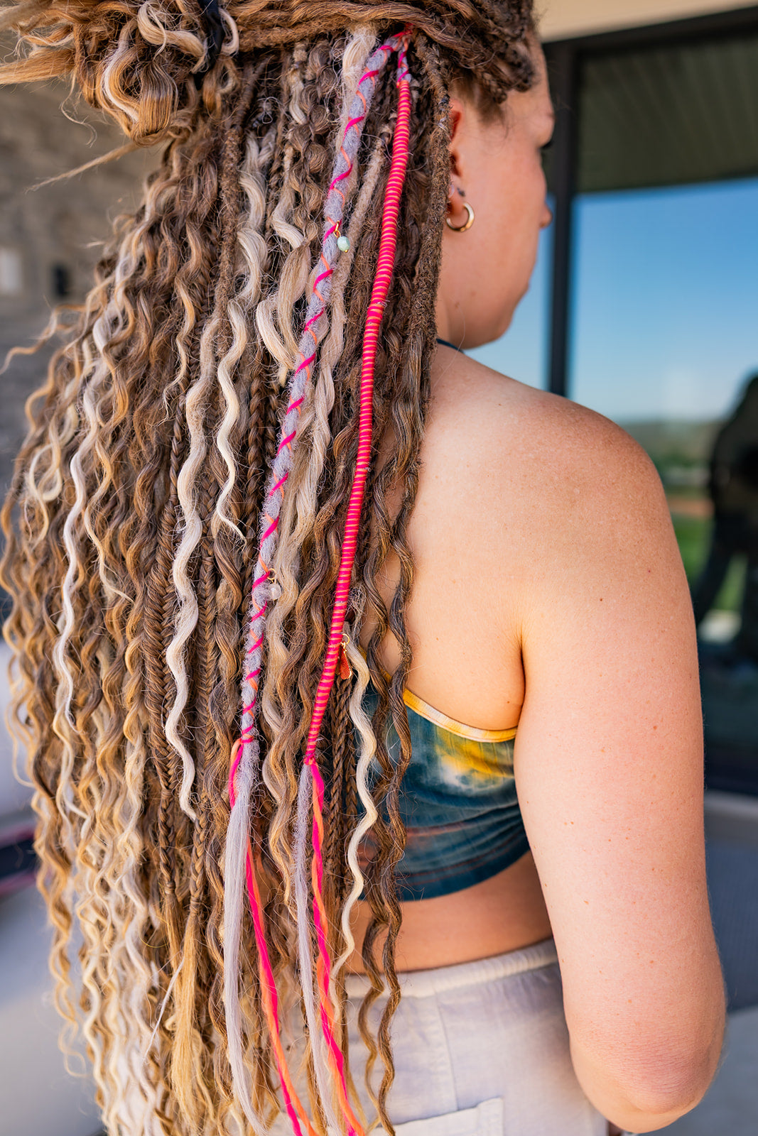 A close-up side view of a person wearing long, wavy dreadlocks with an  accent strand in grey with pink wrap fiber, photographed outdoors near a window.