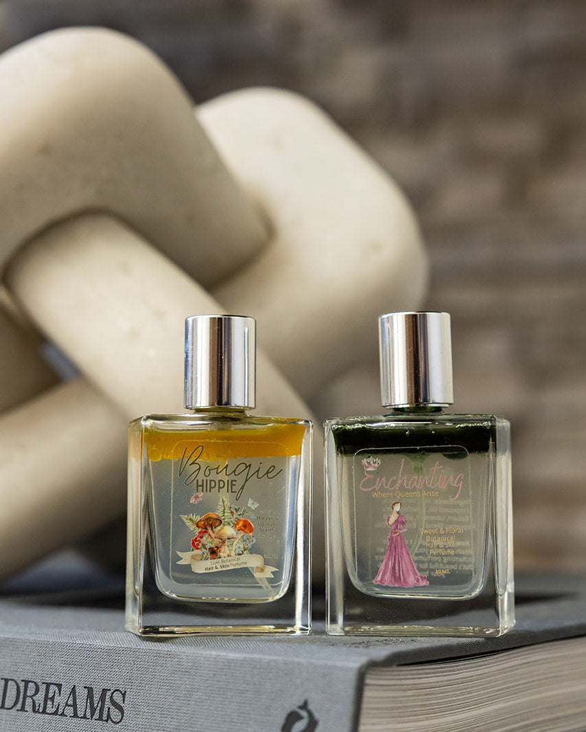 Two small rectangular perfume bottles with silver caps sit on a gray book, styled with a large cream stone knot sculpture and a blurred brick wall background.