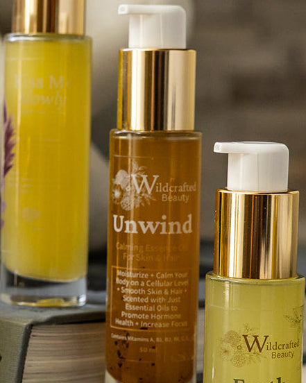 Bottles of gold and amber liquid, focused on the amber body oil in the middle labeled "Unwind"