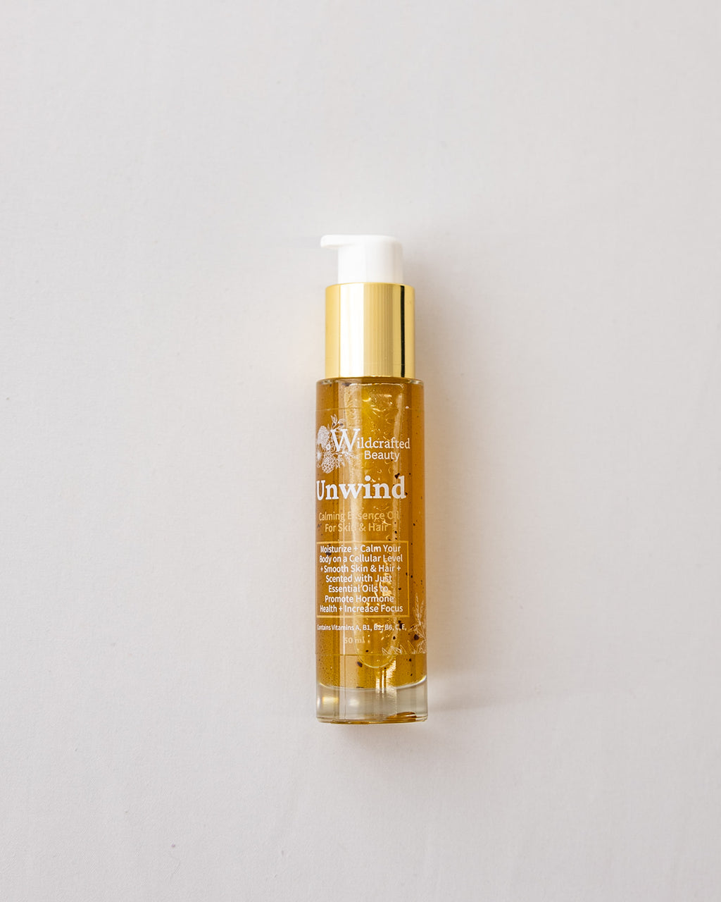 A bottle of Wildrafted Beauty Unwind body oil with golden oil inside, a shiny gold pump collar, and a white pump dispenser, set against a plain light gray background.