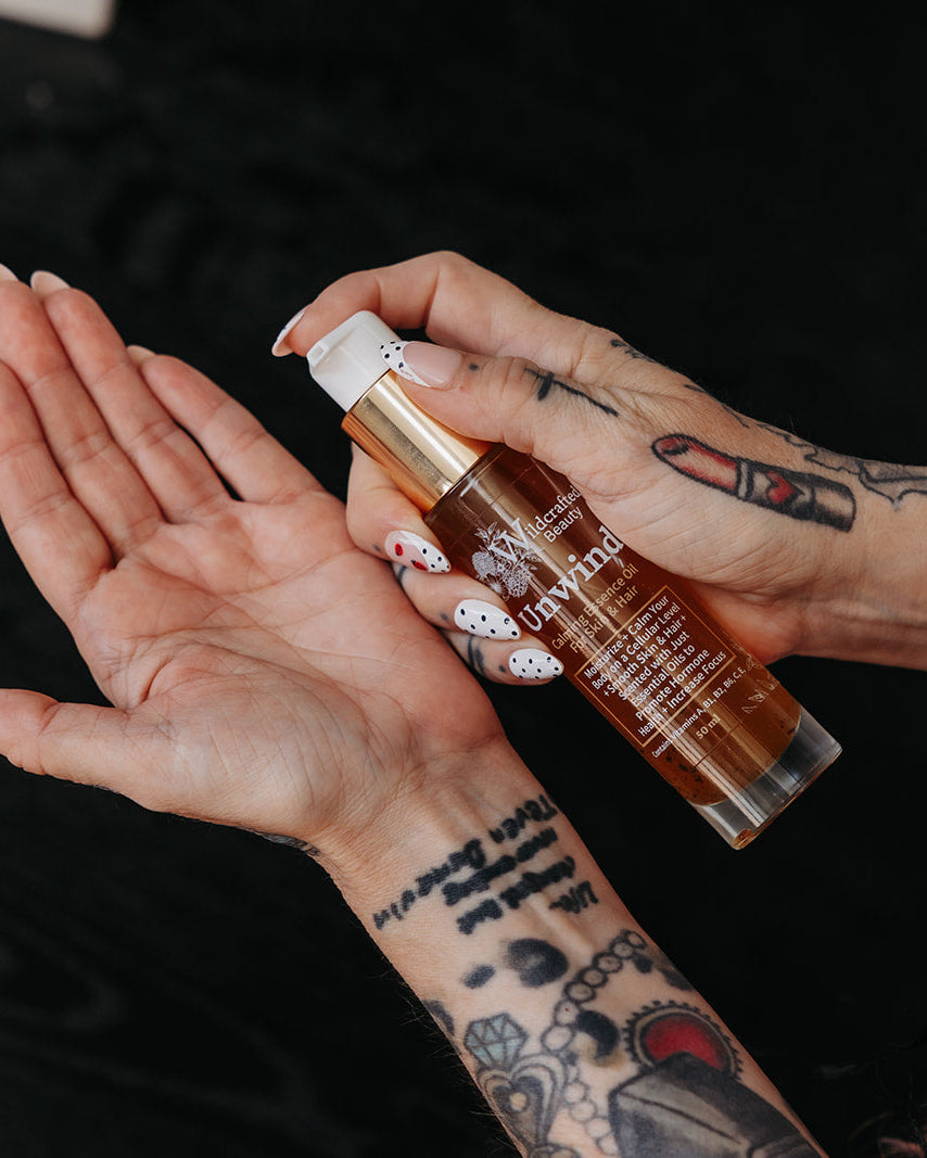 A person with tattooed arms and bracelets holds a clear bottle of amber-colored hair oil over their palm, with the cap being pressed, against a dark background.