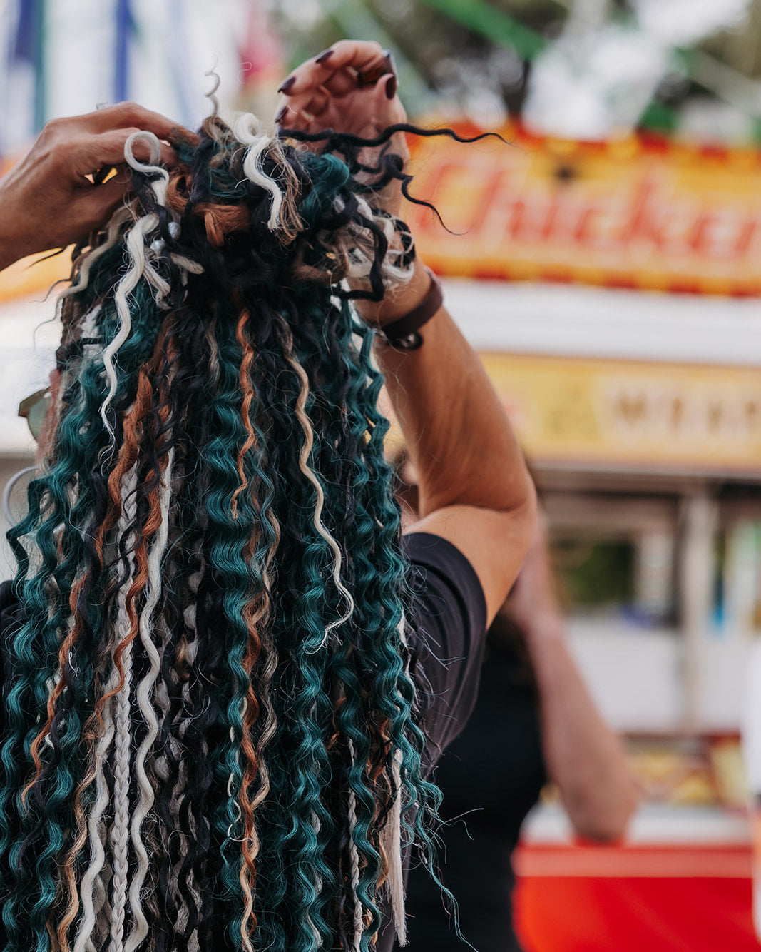 A person at a fair or outdoor event styles long, curly synthetic dreadlocks or hair extensions. The hair is styled in dark teal, black, white, and light brown tones, with a blurred background showing a food stand and another person.