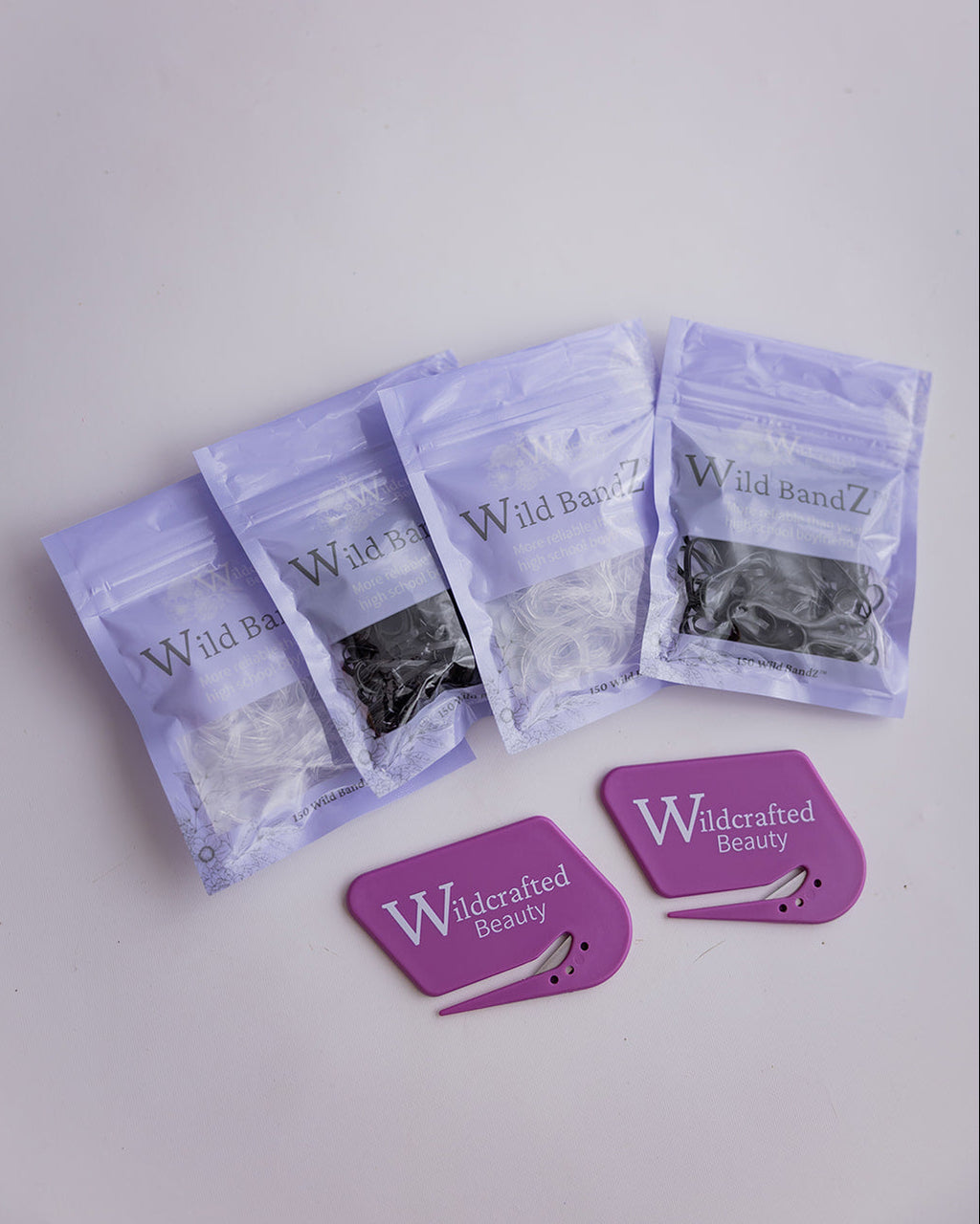 Four clear zip-top pouches containing black or clear hair elastics are arranged on a light gray surface, alongside two purple card-shaped accessories printed with the Wildcrafted Beauty brand.