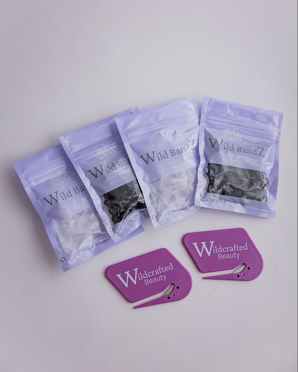 Four clear zip-top pouches containing black or clear hair elastics are arranged on a light gray surface, alongside two purple card-shaped accessories printed with the Wildcrafted Beauty brand.