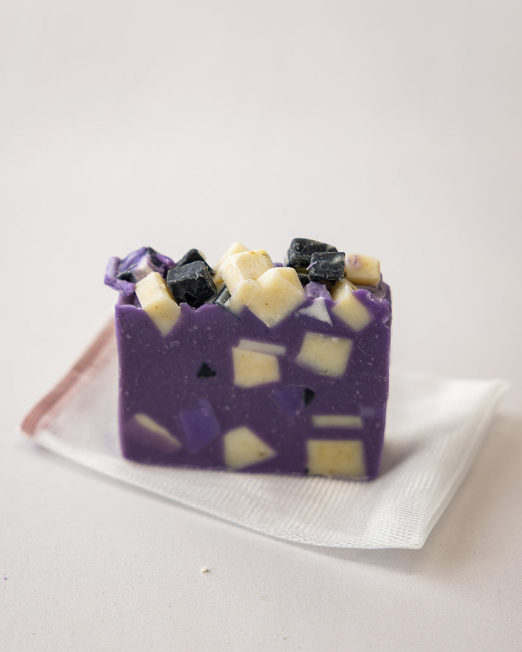 A rectangular bar of purple soap with embedded dark and pale yellow chunks, resting on a white textured paper napkin against a plain light background.