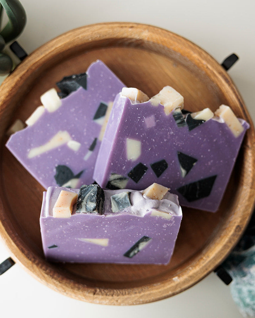 Three rectangular bars of lavender purple artisanal soap with black and white stone inclusions, displayed in a round wooden bowl on a white background with a succulent and a blue-green crystal.