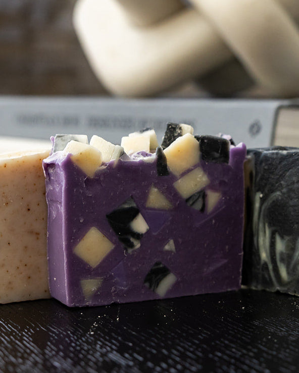 Three artisanal handmade soap bars arranged in a row on a dark wooden surface, featuring purple with cream chunks, black and white marbled, and light beige speckled soap, with a blurred background.
