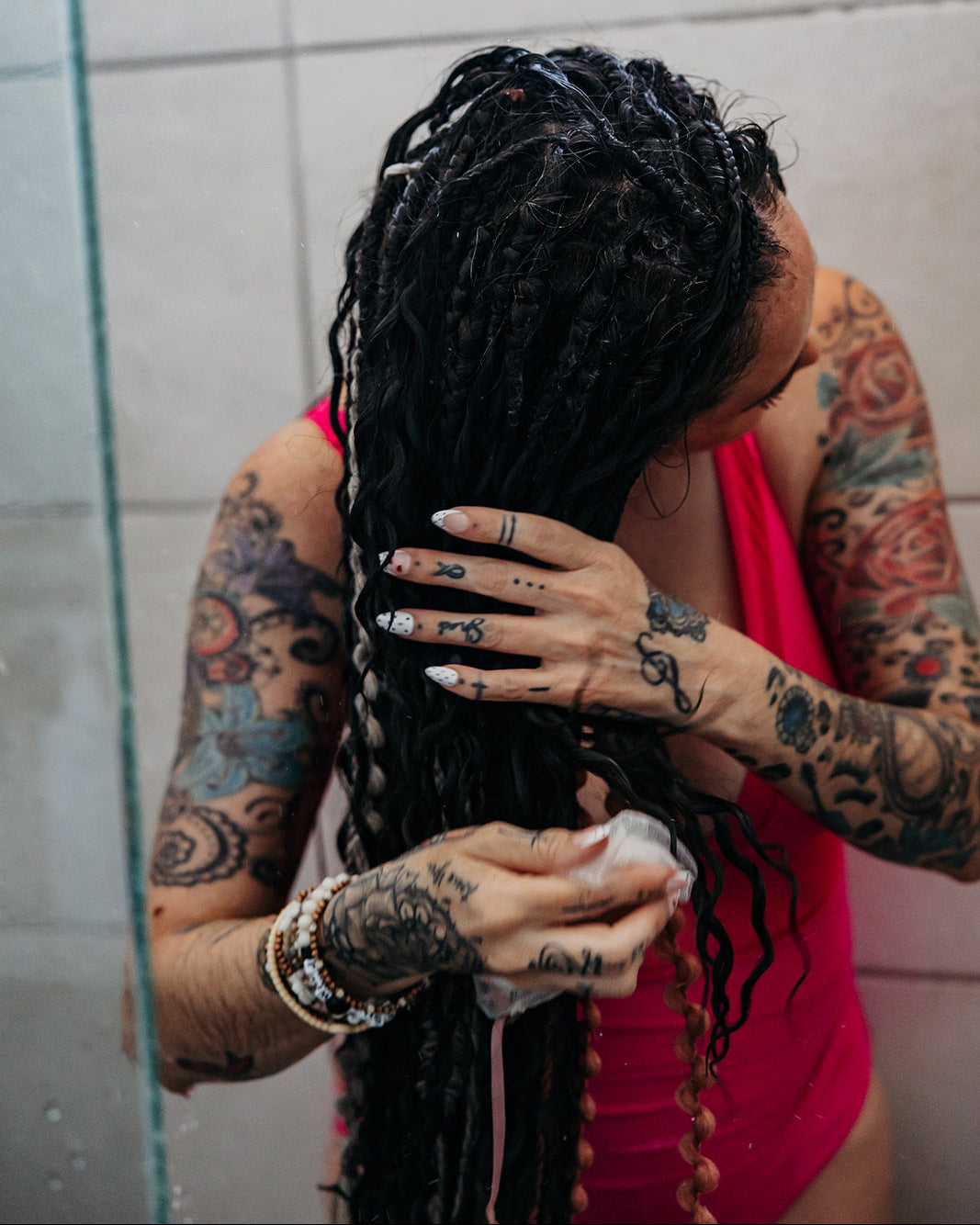 A tattooed woman with long, wet black dreadlocks washes her hair in a shower. She is wearing a bright pink swimsuit top, and a light gray tiled wall is visible in the background.