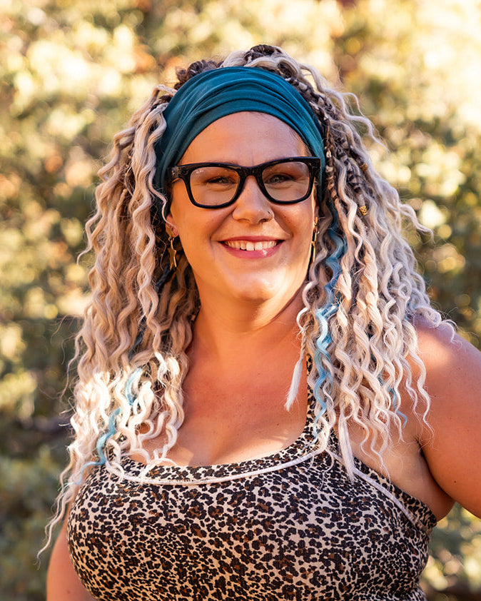 A woman with curly blonde braid-in hair extensions with streaks of blue, wearing glasses, and a teal headband stands outdoors in front of a rocky, sunlit background. She is wearing a leopard-print top.