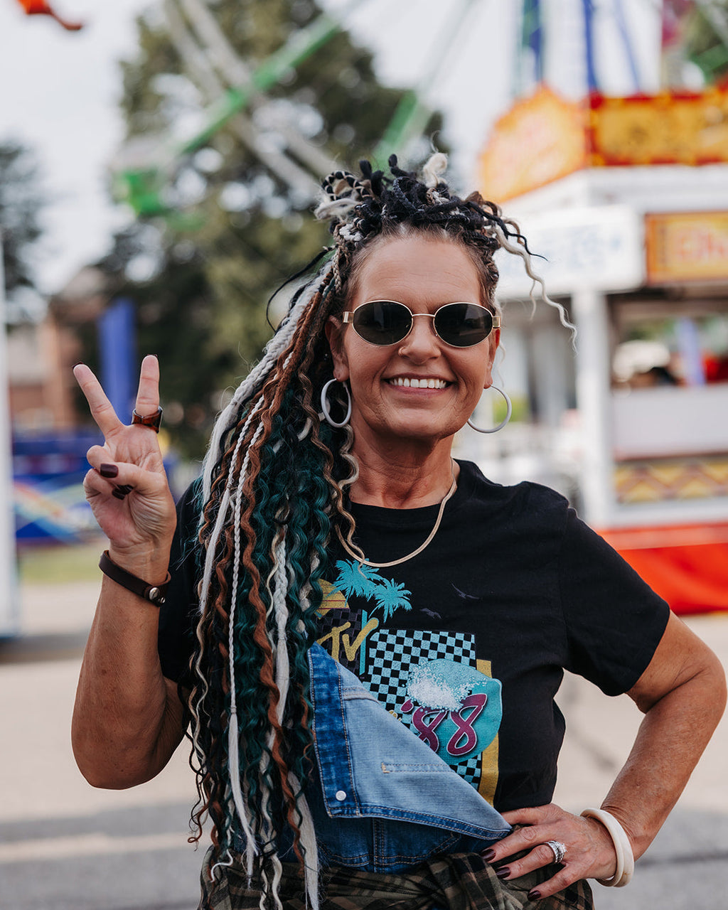 A smiling woman with long braided dreadlocks stands outdoors at what looks like a fairground, wearing round sunglasses, a black graphic T-shirt, a denim jacket tied around her waist, and a plaid shirt. She is making a peace sign with one hand.