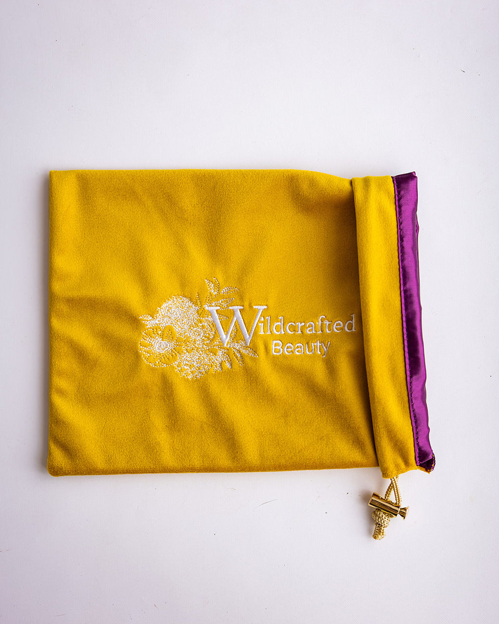 A yellow velvet drawstring pouch with a purple satin inner lining and a white 'Wildcrafted Beauty' logo printed on the front, shown on a plain white background.