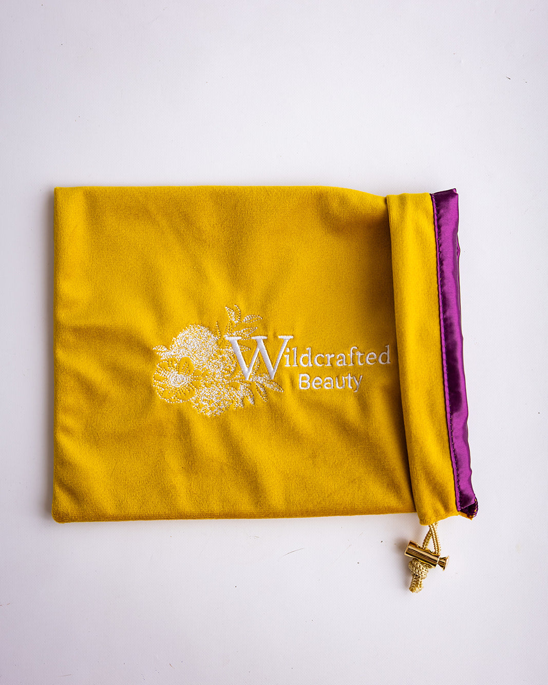 A yellow velvet drawstring pouch with a purple satin inner lining and a white 'Wildcrafted Beauty' logo printed on the front, shown on a plain white background.