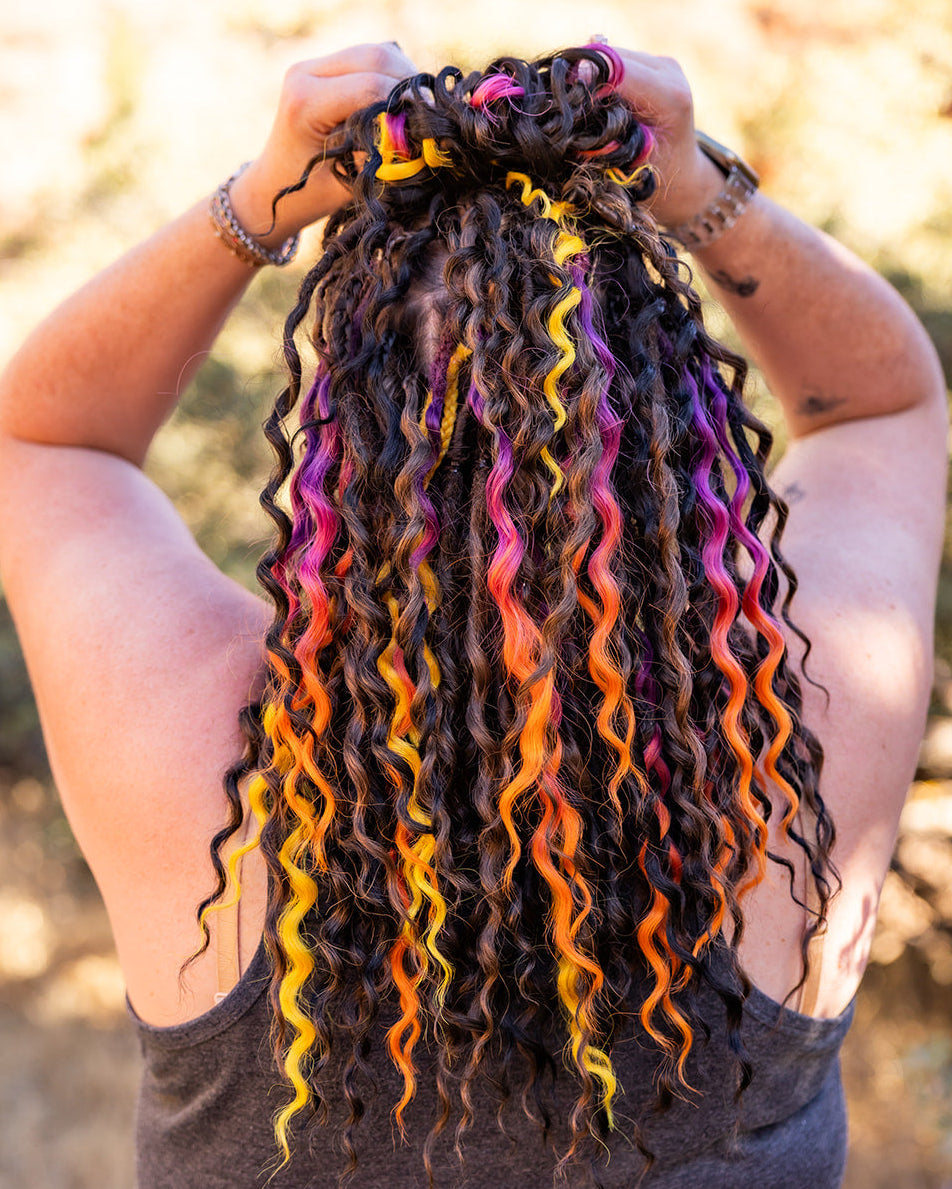 A person outdoors with  curly black braid-in hair extensions highlighted with bright neon streaks of yellow, orange, pink, and purple, seen from behind.