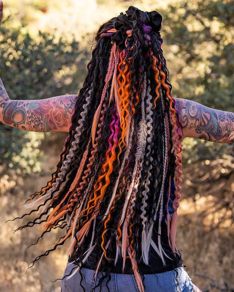 A person outdoors with their back to the camera, wearing a black top and denim shorts, with long, colorful dreadlocks featuring black, orange, pink, and purple tones. Their tattooed arms are raised in a dynamic pose against a blurred natural background of trees and dry grass.